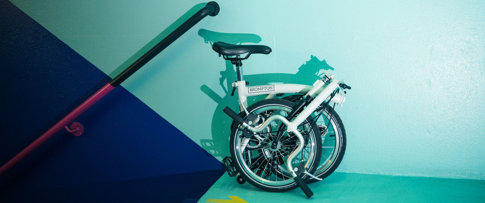 Un Brompton A Line plié contre un mur bleu sarcelle et bleu marine